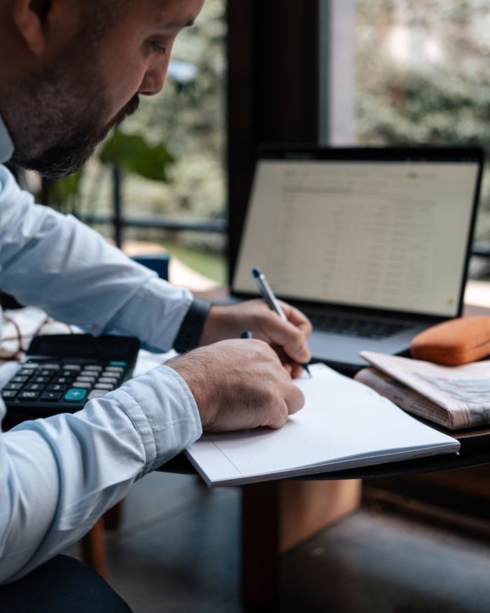 Mastering the First Impression: Your intriguing post title goes here A man writing in a notebook while using a laptop and calculator at a desk.